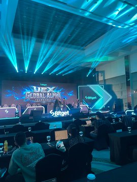 A photo of a USEX Global Alpha event with blue stage lighting, featuring Bitget branding on the stage and screens in a large venue with attendees seated at tables.