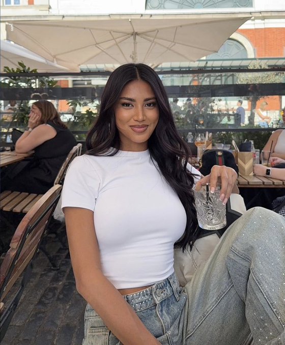 A young woman with long dark hair wearing a white fitted t-shirt and denim jeans, sitting at an outdoor café holding a glass.