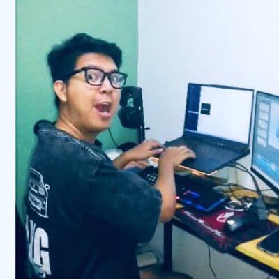 A young man wearing glasses and a dark t-shirt sits at a desk with multiple monitors, typing on a keyboard with an excited, open-mouthed expression.