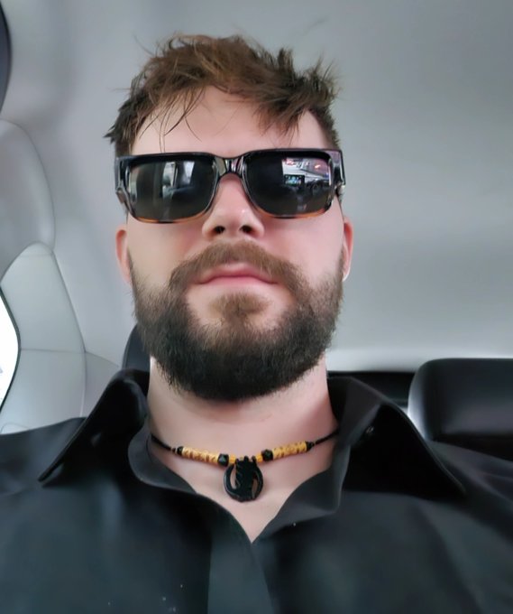 A man with a thick beard and sunglasses sits inside a car, wearing a black shirt and a distinctive necklace with yellow beads and a dark pendant.