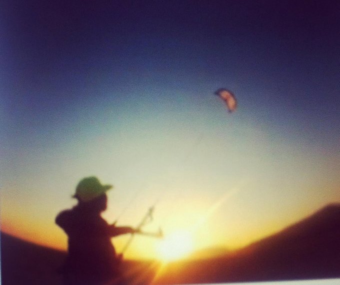 A silhouetted figure wearing a hat flies a kite-shaped parachute against a vivid sunset sky with mountains in the background.