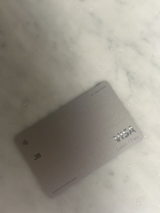 A gray metallic Visa Debit/Flex card with 'JB' engraved on it, resting on a white marble surface.