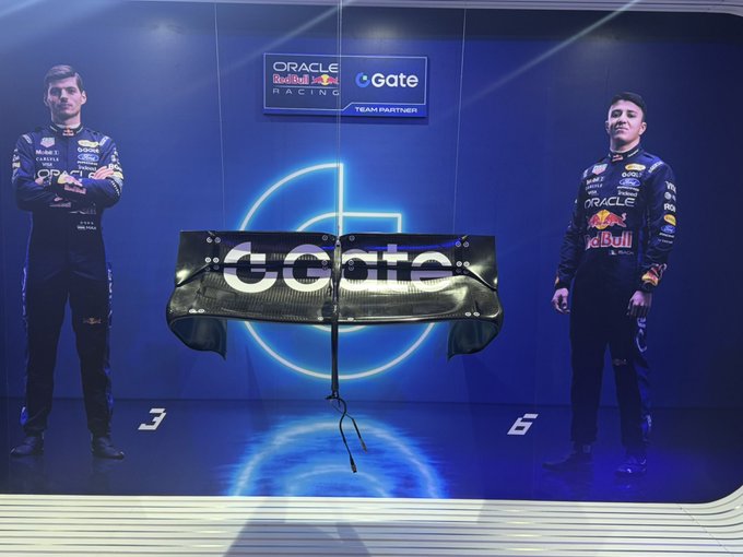 Two Red Bull Racing drivers in Oracle team suits stand on either side of a rear wing displayed with the 'Gate' logo at a team partner event.
