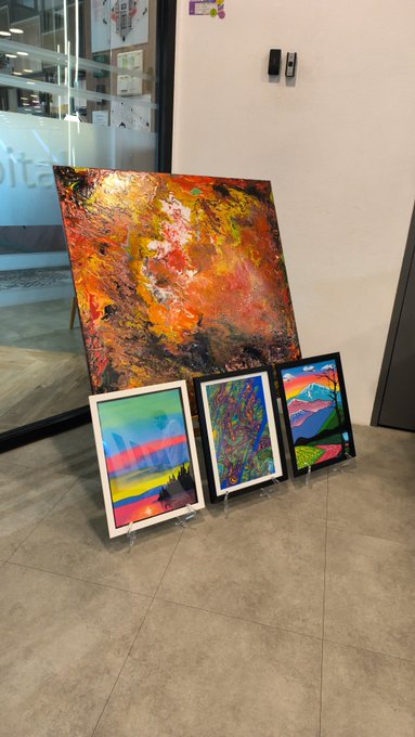 A display of four abstract paintings arranged on a tiled floor in what appears to be a lobby, with a large textured, warm-toned painting leaning against the wall and three smaller framed pieces propped in front.