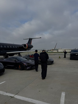 A private jet is parked on an airport tarmac near a sleek sports car, with several people standing nearby under a cloudy sky.