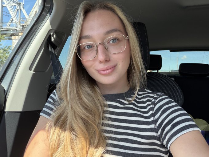 A young woman with long blonde hair and glasses takes a selfie in her car, wearing a black and white striped shirt. The relaxed, casual photo complements the laid-back tone of her post asking about "$Pistacio."