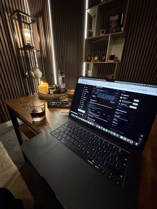 A laptop on a wooden desk displaying code, with a glass of dark liquid, a candle, and a bookshelf visible in a dimly lit room.