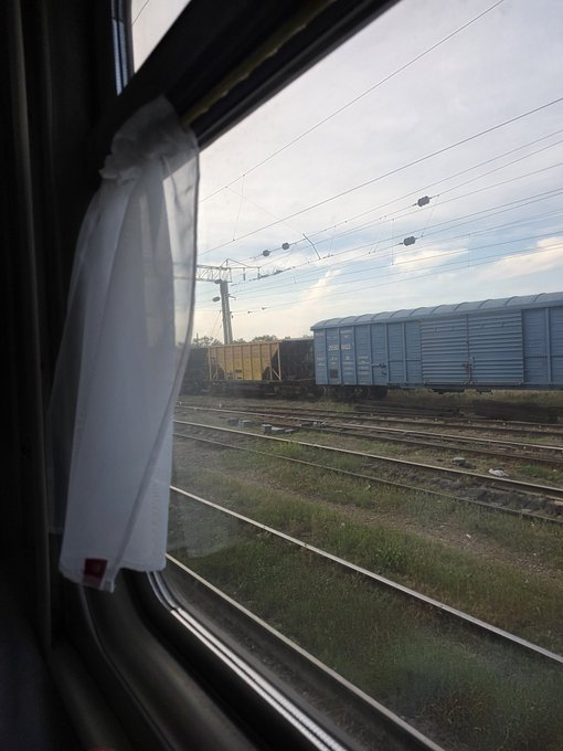 The image shows a train window with white curtains that are open, providing a view of an outdoor scene outside. The train tracks stretch out into the distance, leading towards another train further away from the viewer's perspective. In addition to the trains, there is also a car visible in the background. The post includes a link to a football game and mentions that someone has bet on it.