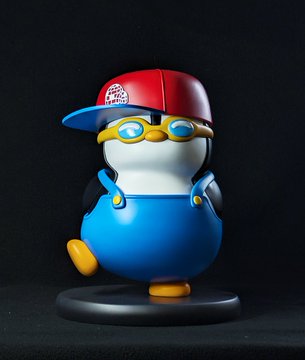 A 3D rendered penguin figurine wearing a red and blue cap, blue sunglasses, and blue overalls, posed on a small black base against a dark background.