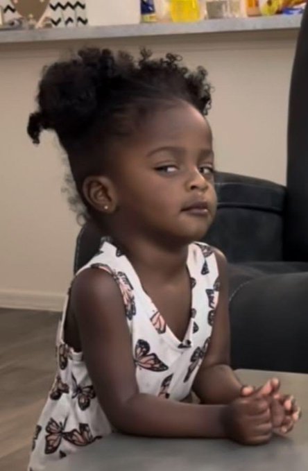 A young girl in a butterfly-print outfit sitting at a table with a skeptical or unimpressed facial expression.