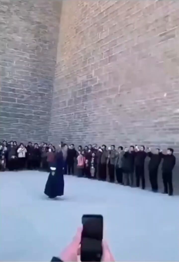 urns of water are visible through a stone wall, with people gathered on the right side and a person holding a phone on the left.