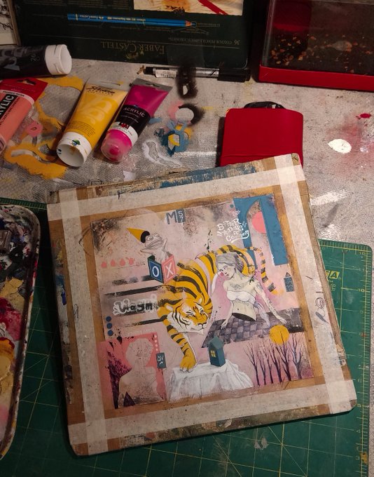 An artist's workspace featuring a mixed-media collage artwork with a tiger, figures, and text like 'YES' and 'Sunshine', surrounded by paint tubes on a green cutting mat.