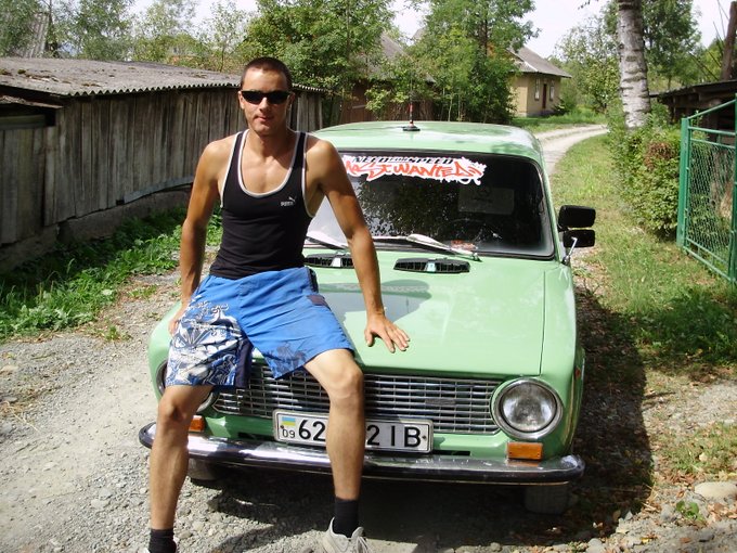 The image features a man posing next to an old green car parked on a dirt road. The man is wearing blue shorts and sunglasses while leaning against the car with his arms resting on its hood. The photo has been shared, and the caption reads "It's my birthday today. Found this old photo and it made me smile a bit. A lot has changed since then - life, markets, people, everything. But I'm still here, still learning, still building."