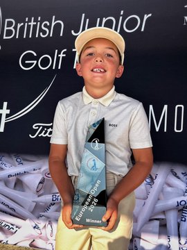 A young boy, smiling proudly while wearing a white cap and Hugo Boss polo shirt, holds a "British Junior Golf Tour Winner" trophy at the European Open event, celebrating his victory in the competition.