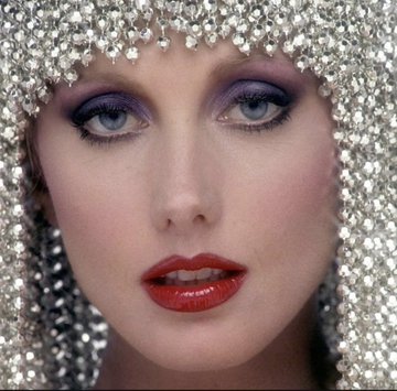 A close-up portrait of a woman with striking blue eyes, purple eyeshadow, and red lipstick, framed by a beaded silver headpiece.
