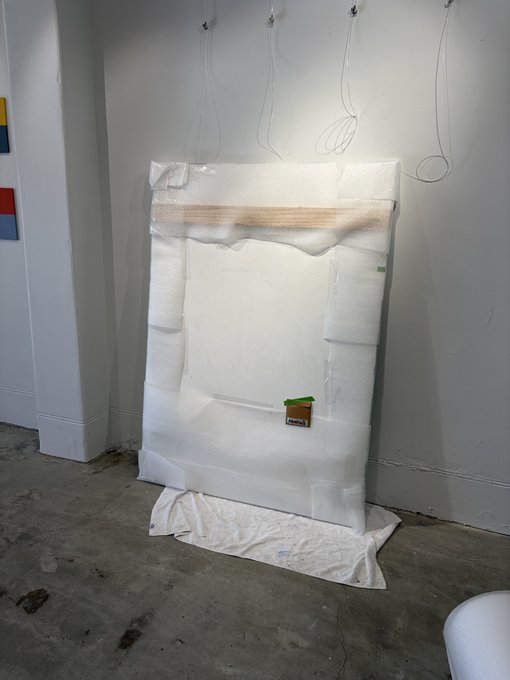 A large canvas painting wrapped in protective bubble wrap and cloth leans against a gallery wall, displayed without being hung up. The image humorously illustrates the caption's sentiment—highlighting the irony of an artwork sitting uninstalled on the floor, ready to be sold to any collector rather than limited to NFT buyers.