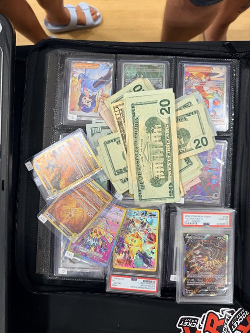 An open black binder displaying graded Pokémon cards in plastic slabs with cash (US $20 bills) placed on top, with a person's feet in sandals visible in the background.