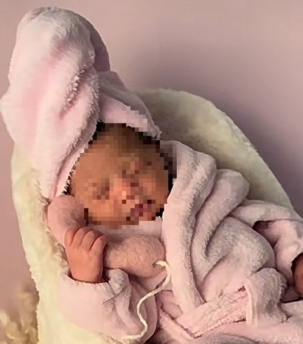 A newborn baby wrapped in a soft pink hooded blanket, with their face pixelated for privacy, lying against a pale background.