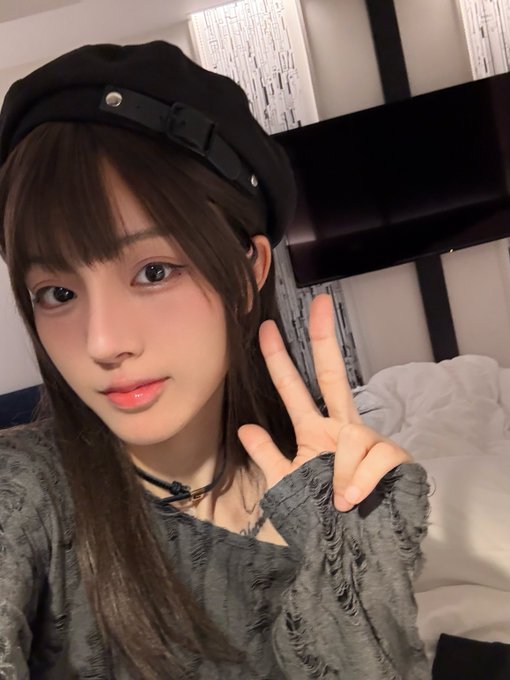 A young woman wearing a black beret and gray textured top flashes a peace sign, posing in what appears to be a hotel room setting. The image accompanies a promotional post inviting friends to join OKX prediction matches for a chance to earn 100XP through simple participation steps.
