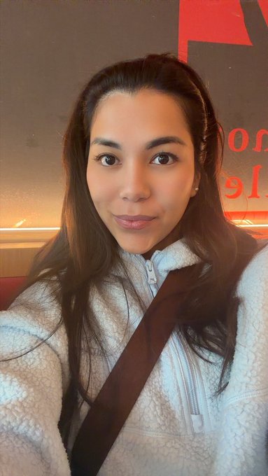 A woman with long brown hair wearing a fluffy cream-colored fleece jacket and a brown crossbody bag strap takes a selfie.