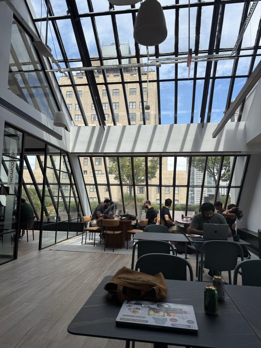 A bright, modern café or common area with large angular windows overlooking other buildings, where several people are seated at tables working on laptops.