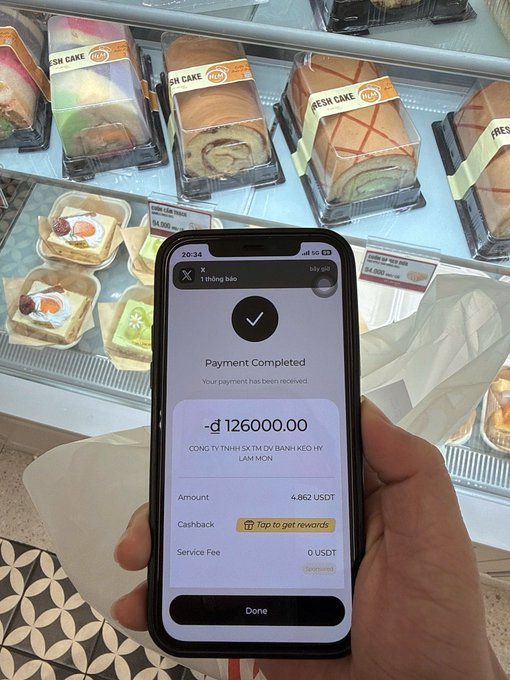 The image shows a person holding up their smartphone displaying a digital payment method, possibly from Stone Wallet. The screen of the phone reveals an account balance of $12,000. The text on the photo also mentions that the user has paid for something in the USA and received an amount to be paid back.