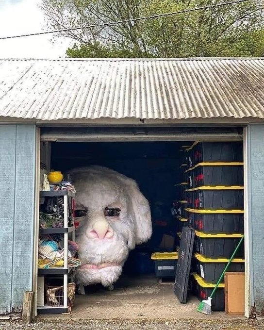 A person wearing a fluffy gray monster costume with a pink nose stands inside a shed filled with stacked storage bins, tools, and shelving.