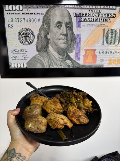 A hand holds a black bowl of food containing chicken and vegetables beneath a framed $100 US bill (Federal Reserve Note) mounted on a wall.