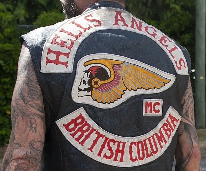 A person wearing a black leather vest displays the 'HELLS ANGELS MC BRITISH COLUMBIA' back patch, featuring the iconic winged skull logo, with visible tattoos on their arms.