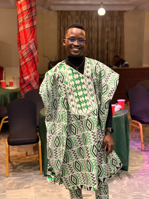 A smiling young man wearing glasses, a black turtleneck, and a green and white patterned traditional African outfit, standing in an event hall.