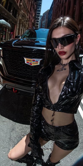 The image features a woman wearing black leather clothing, including shorts and boots, posing next to a Cadillac Escalade car with its hood open. She has her legs crossed and is kneeling down, giving off an edgy and stylish vibe. The caption "QT your ride" suggests that the woman might be promoting or discussing the car in some way.

The image also includes text elements such as "QT your ride", which could potentially be related to a promotional campaign for the Cadillac Escalade. However, without more context or information about the content of the caption, it is difficult to determine its exact connection to the image.