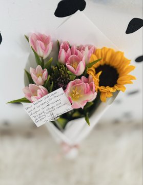 A small bouquet of pink and yellow flowers wrapped in white paper with a handwritten note attached.
