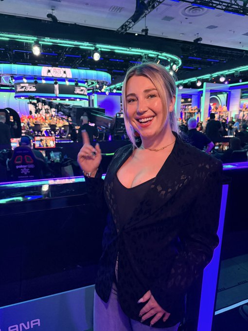 A smiling woman with blonde hair wearing a black top and patterned cardigan, pointing upward at what appears to be a poker tournament event.