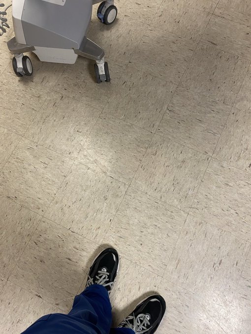 A downward view shows black sneakers on a person standing on a speckled linoleum floor, near the wheels of a gray medical cart.