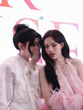 Two women pose closely together at what appears to be a fashion event, with one wearing a white feathered blouse and the other a pink halter dress with feather trim.