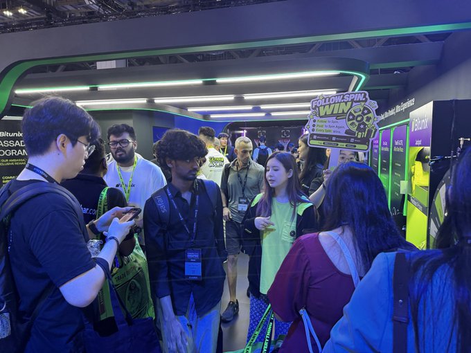 A bustling crowd gathers around a "Follow Spin to Win" promotional booth at what appears to be the Token2049 crypto conference, with attendees wearing lanyards and badges as they wait their turn. The image captures the curious, slightly overwhelmed feeling of a first-time visitor surveying the diverse mix of people drawn to the event.

Visible signage includes "Bitunix," "ASSAD," "KASSA," and "Follow Spin to Win," suggesting promotional activity from various cryptocurrency exchanges and projects.