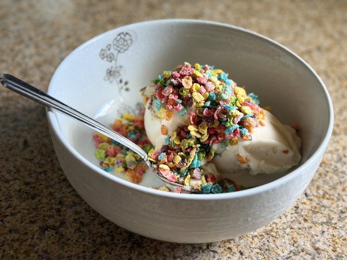A white bowl filled with scoops of ice cream or yogurt generously topped with colorful, fruity cereal pieces, with a spoon resting inside.
