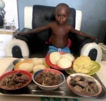A young boy sits shirtless at a table with a large tray of food in front of him, including what appears to be fufu, rice, and various meat dishes.