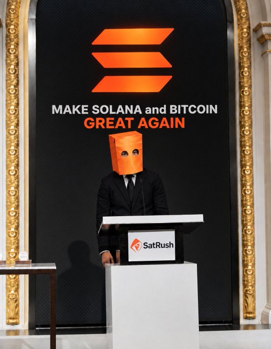 A person in a suit wearing an orange paper bag over their head stands at a podium labeled 'SatRush' in front of a sign reading 'MAKE SOLANA and BITCOIN GREAT AGAIN'.