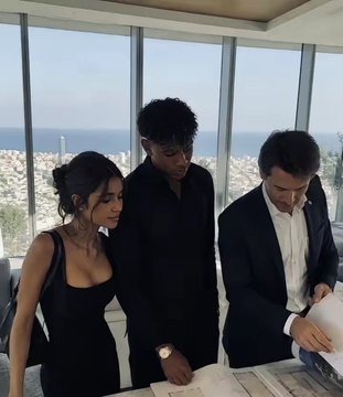 Three people in formal black attire stand at a high-rise window overlooking a city and ocean, examining documents on a table.