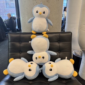 Four plush penguin toys are arranged on a black leather chair; one sits upright on top of the chair back while three lie stacked together on the seat.