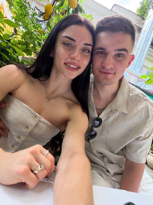 A close-up selfie of a smiling young couple, with the woman wearing a strapless beige top and a ring on her finger, and the man in a tan shirt with sunglasses hanging from the collar, posed in front of a lemon tree.