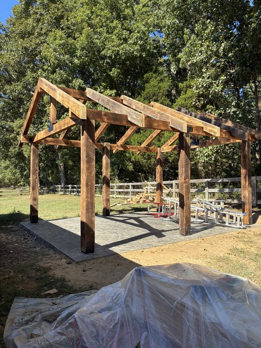 A wooden pavilion or pergola structure under construction in a grassy outdoor area.