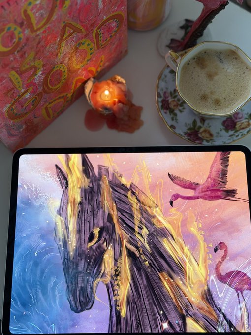 A digital drawing of a horse engulfed in flames alongside pink flamingos displayed on a tablet, surrounded by lit candles, a floral teacup, and a red decorative box.
