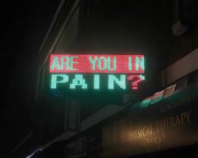 A nighttime photograph of an illuminated LED sign reading "ARE YOU IN PAIN?" in red and green text, mounted above a physical therapy office.
