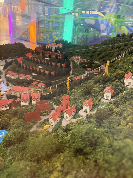 Close-up photo of a detailed miniature model town with small houses featuring red roofs nestled in green model trees and terrain, with colorful structures visible in the background.