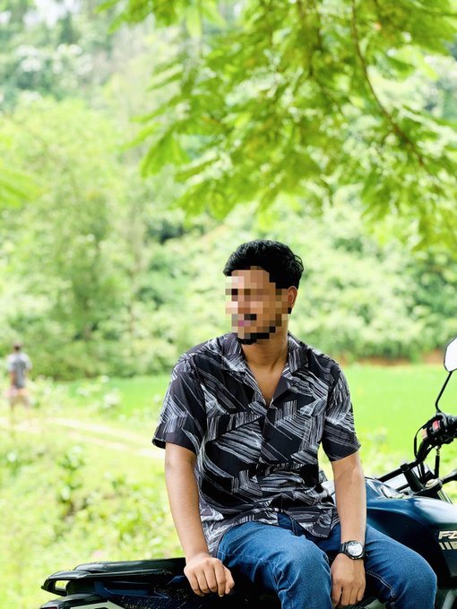 A man with a blurred face wearing a patterned shirt sits on a motorcycle in a lush green outdoor setting.