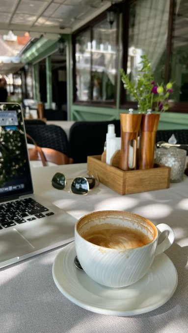 A coffee cup with latte on a saucer sits on a cafe table beside a laptop, sunglasses, and a caddy with condiments and flowers.