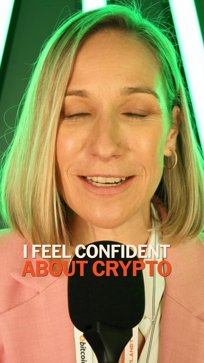 The image features a woman with blonde hair wearing a pink blazer, smiling at the camera while holding a microphone. The background of the image has neon green lights that are turned off, making her stand out against the dark backdrop. Below the image, there is text that reads "I feel confident about crypto". This suggests that the woman in the image might be an advocate for or supporter of cryptocurrency and its benefits.