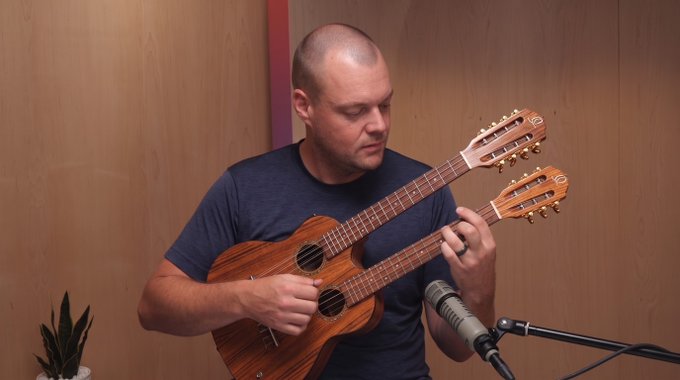 A man plays a unique double-necked ukulele in front of a microphone, with wood paneling behind him and a small plant visible to the side. The post about "Gemini dropping tomorrow" appears unrelated to the image content.